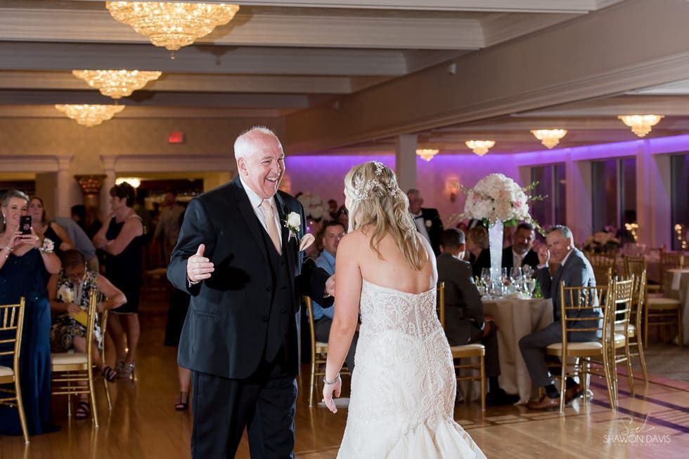 Bride in white dress dances with older man in black suit at a formal reception hall