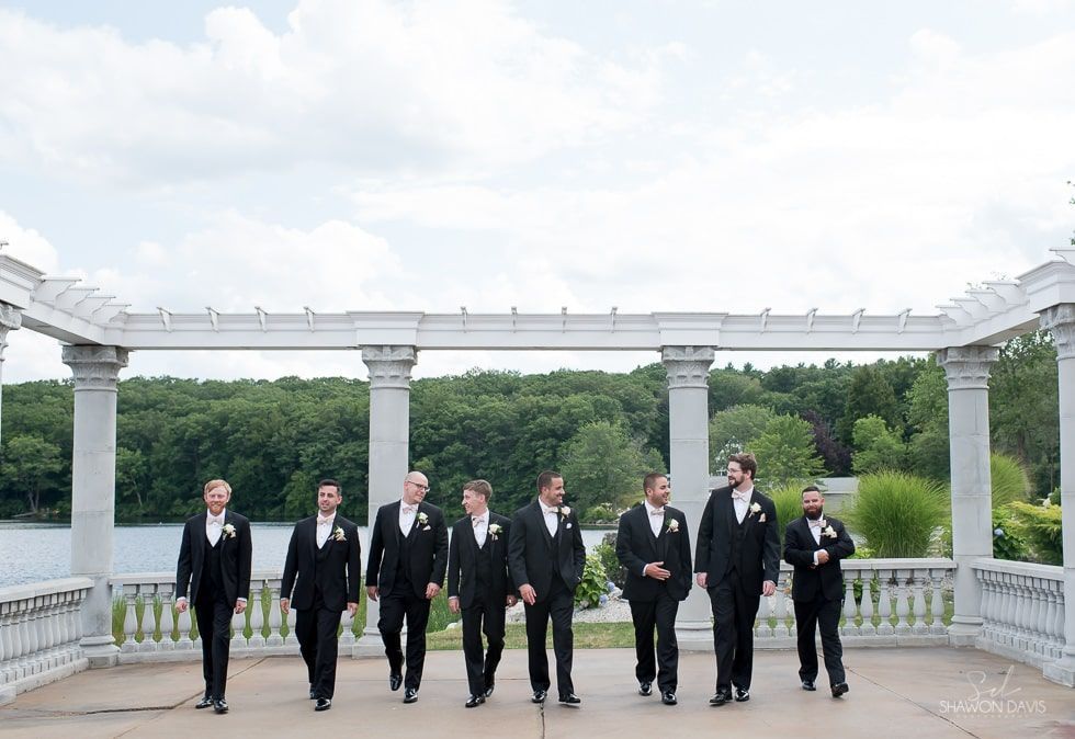 Seven people in black suits stand beneath white columns on a balcony overlooking a green landscape.
