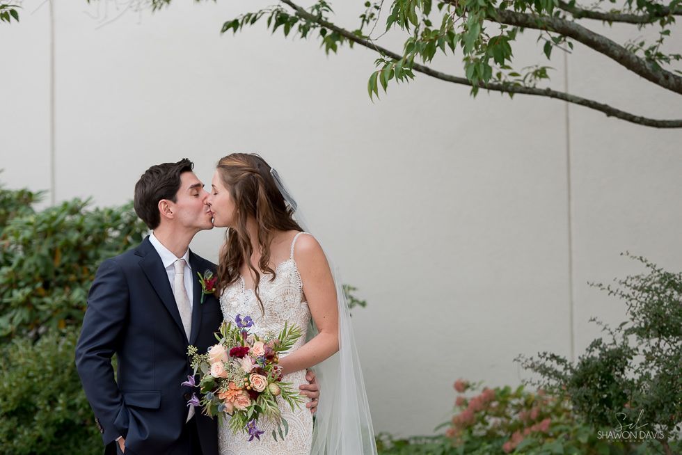 Newlywed couple kissing outdoors, bride holding a colorful bouquet by a white wall