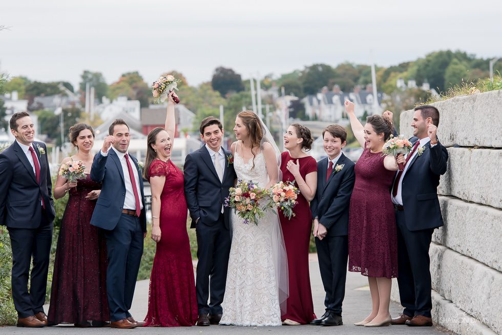 Wedding party posing outdoors by a stone railing, with the bride and groom centered among guests holding bouquets.