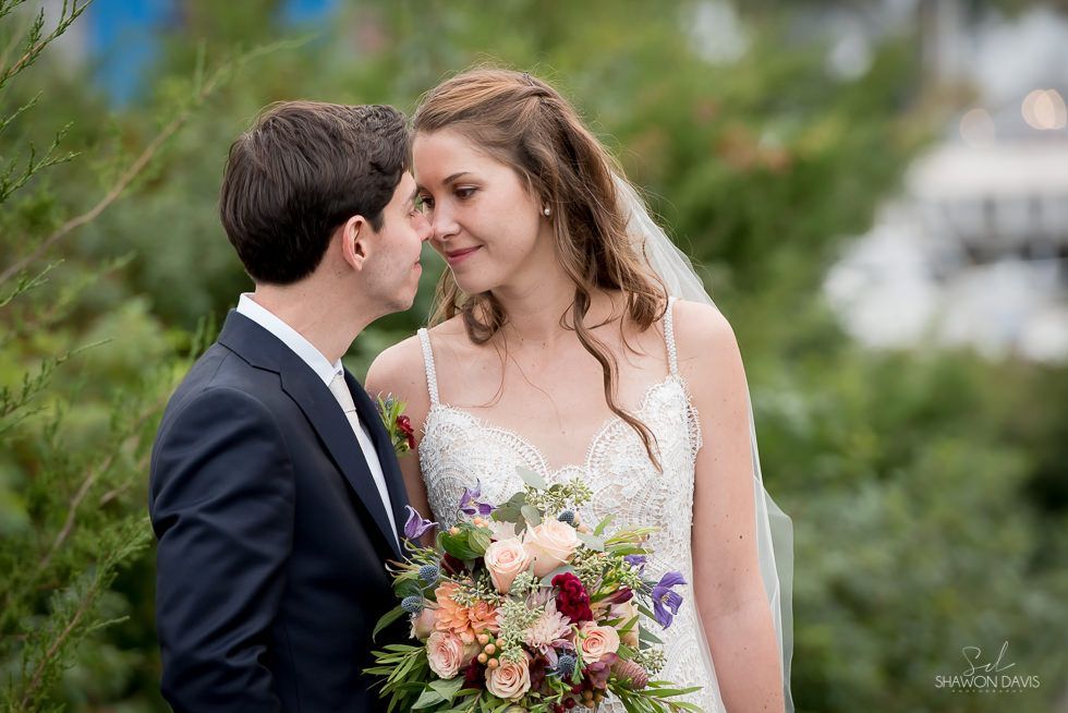 Bride and groom smiling outdoors, holding a bouquet amid greenery.