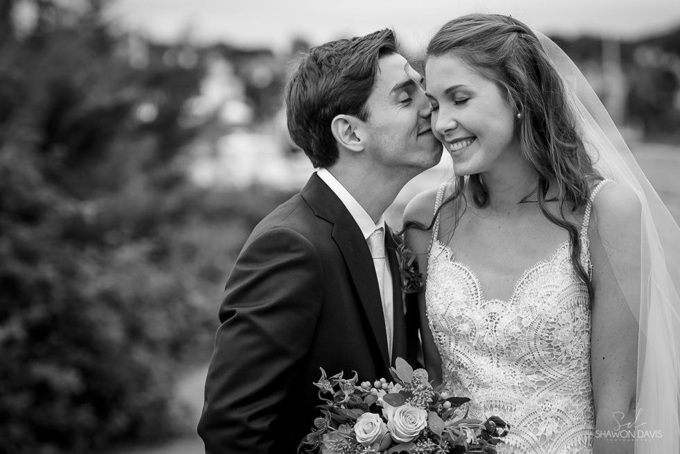 Bride and groom smiling outdoors, with the groom kissing the bride on the cheek.