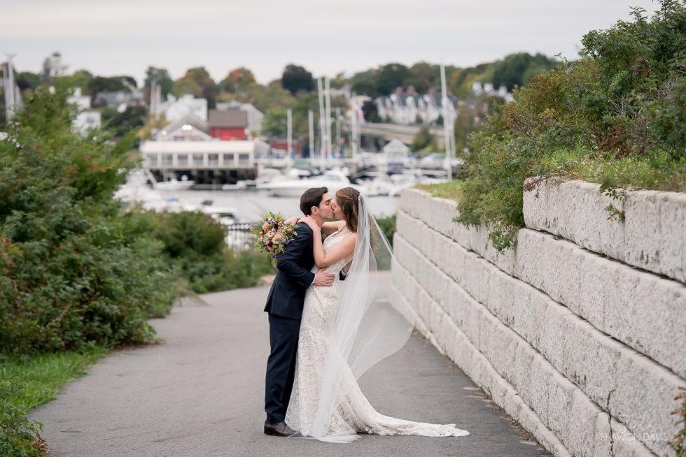 Bride and groom kiss beside a stone wall with a marina and trees in the background