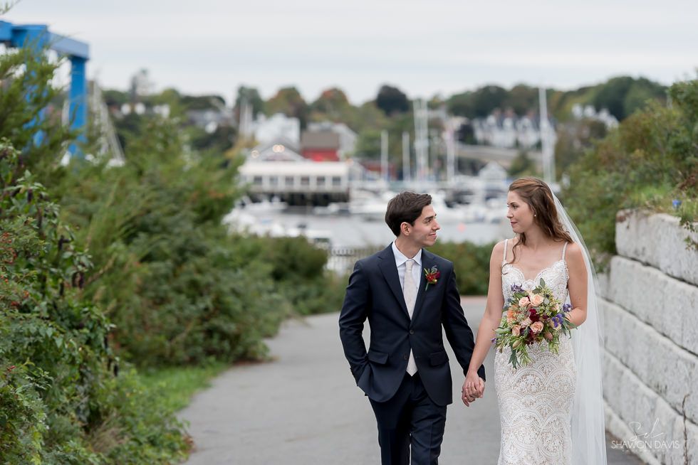 Bride and groom holding hands and smiling on a waterfront path near boats and greenery