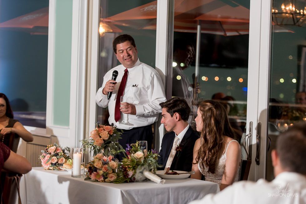 Speaker at a wedding reception toast beside a flower-decorated table, with guests seated nearby