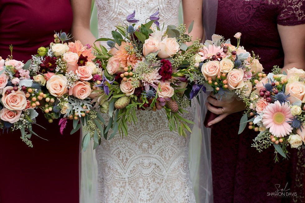 Bride holds a cascading bouquet of pale pink, peach, and purple flowers against a lace wedding dress