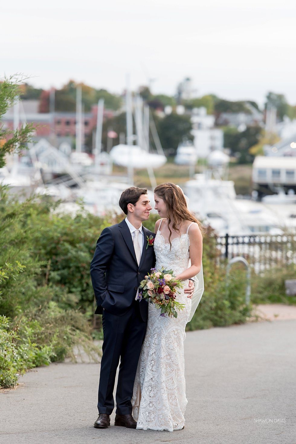 Couple in wedding attire embracing on a path by a harbor with boats in the background