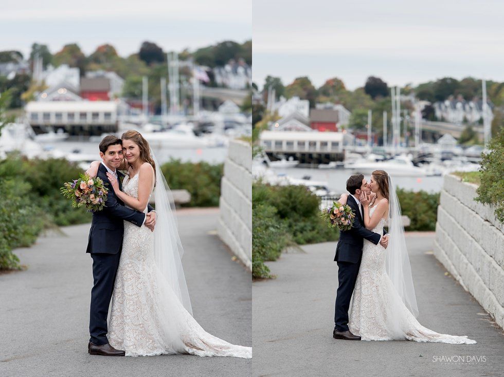 Bride and groom embrace on a waterfront walkway with boats and houses in the background.