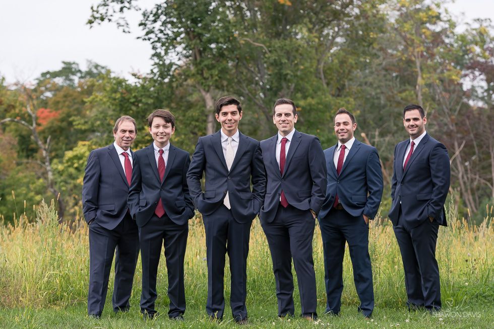 Six men in dark suits and red ties standing together outdoors on grass with trees behind them