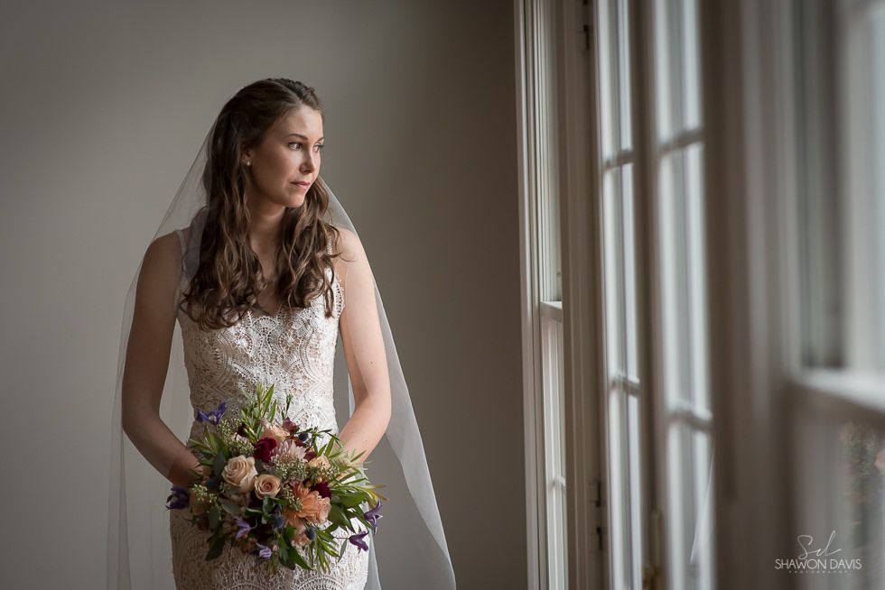 Bride in a white lace dress holding a colorful bouquet by a window.