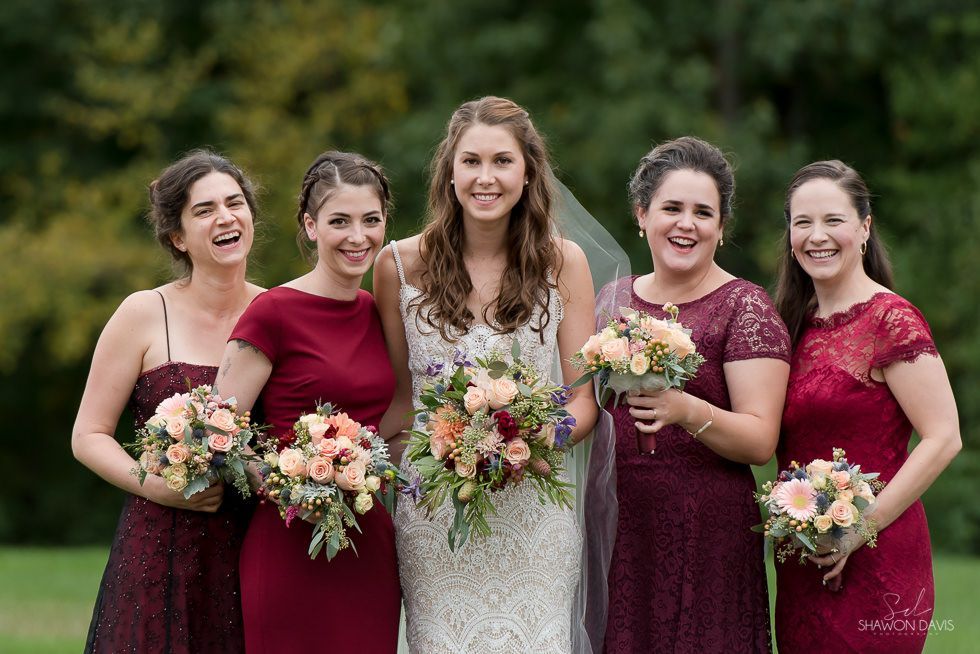 Five women in burgundy dresses and a white gown, holding bouquets outdoors on a grassy path.