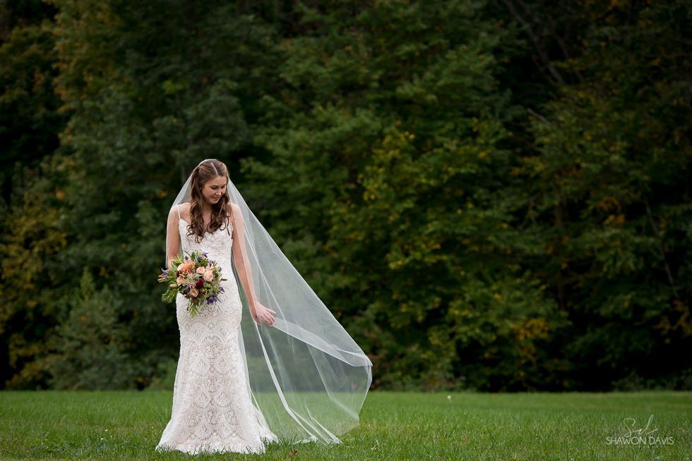 Bride in a white lace gown holding a bouquet on a grassy field with a long veil, against a forest backdrop