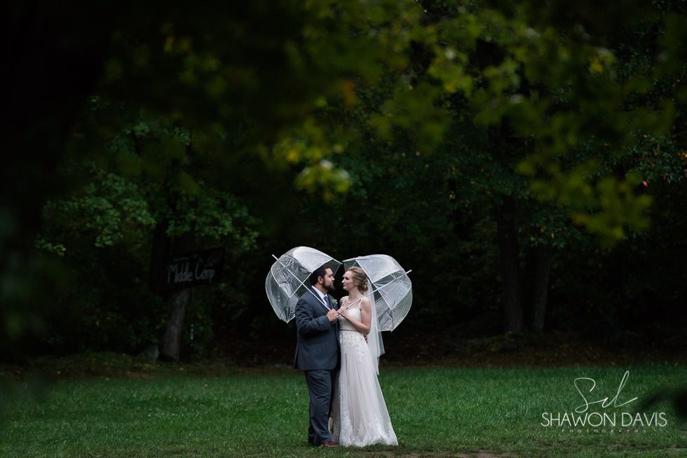 Bride and groom under a clear umbrella in a dark green garden, sharing a close embrace.