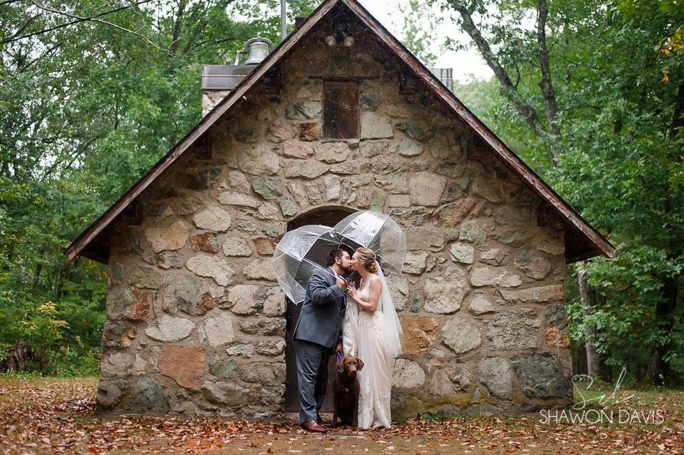 Couple standing under a clear umbrella in front of a stone chapel in a wooded setting