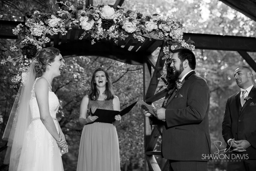 Outdoor wedding ceremony under a floral arch, bride and groom facing officiant with guests nearby