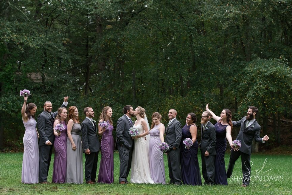 Wedding party posing outdoors in a wooded area, with bridesmaids in lavender and groomsmen in dark suits.