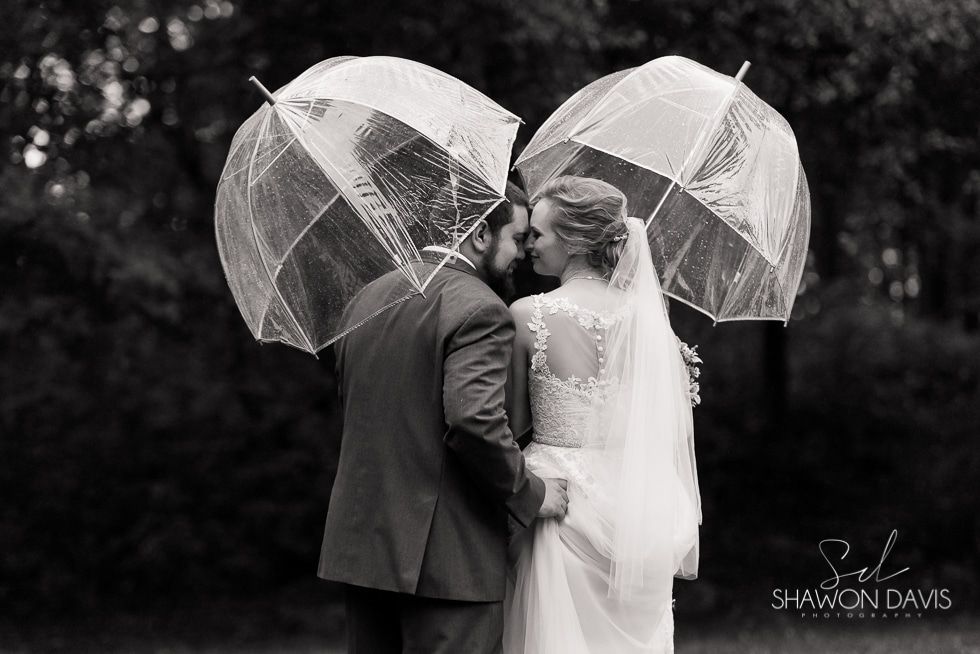 Bride and groom kiss under clear umbrellas in a black-and-white outdoor wedding scene