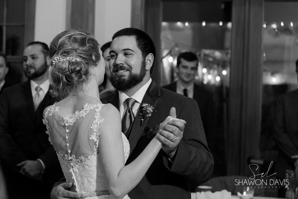 Couple slow dancing at a wedding reception, smiling and holding each other in a dimly lit room.