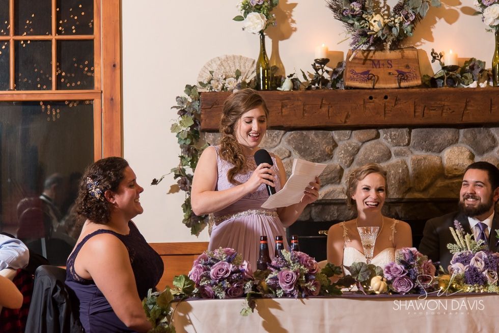 Woman giving a speech at a decorated indoor reception table with two seated guests and a stone fireplace behind her