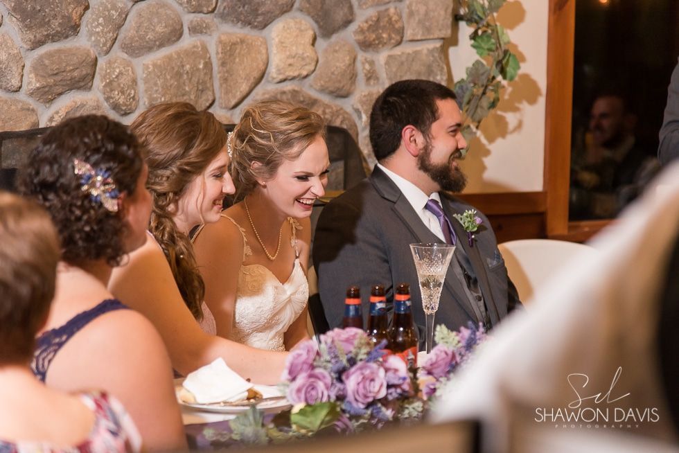 Bride and groom laughing at a wedding reception table with purple flowers and drinks