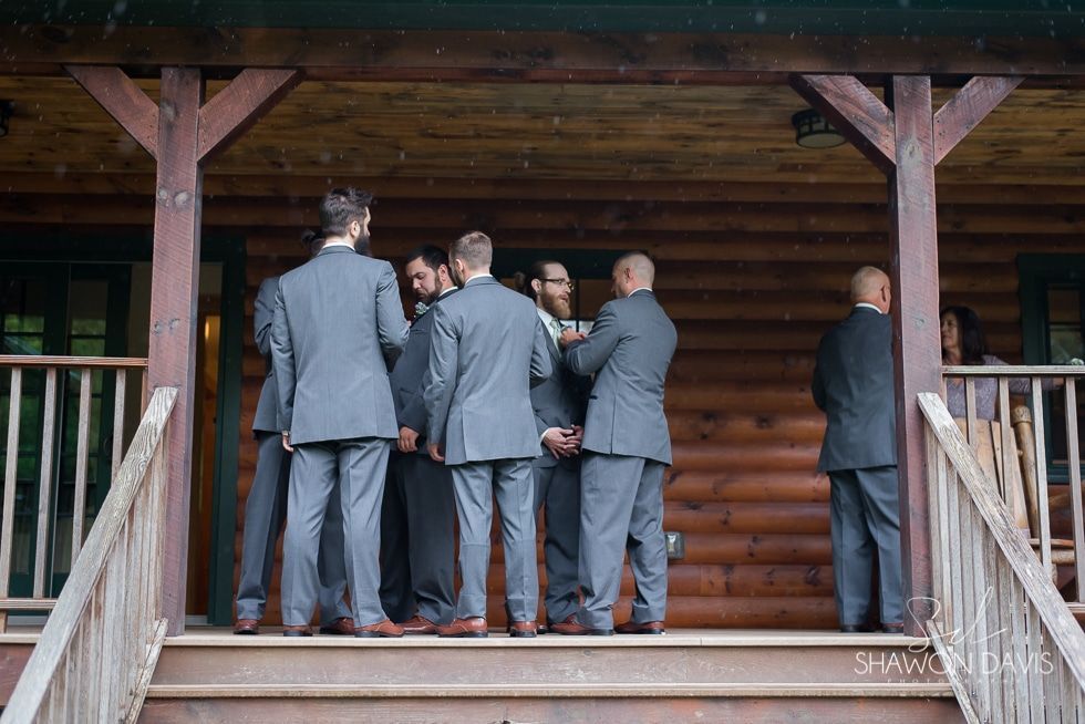 A group of men in suits stands on a wooden porch under a log cabin roof, facing inward in conversation.