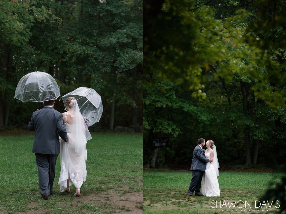 Two couples in wedding attire walk under clear umbrellas in a wooded park.