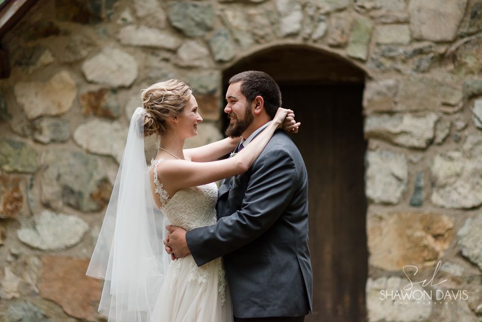 Bride and groom embracing in front of a stone wall, wedding veil flowing