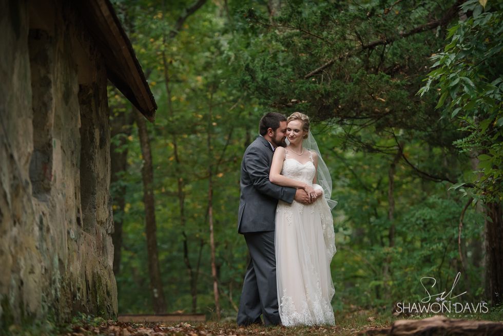 Bride and groom embrace in a forest beside a rustic stone wall.