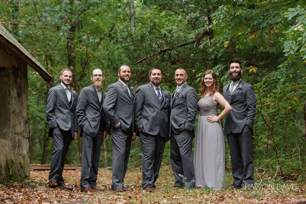 Seven people in formal attire posing outdoors in front of trees and a stone building.