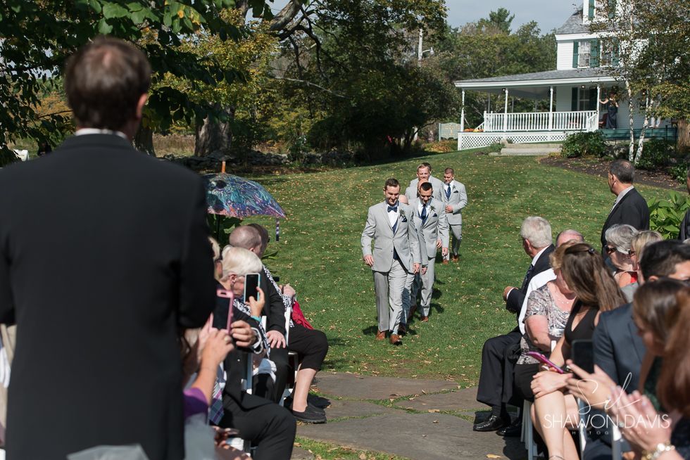 People seated outdoors watch a procession of people in gray suits walking down a garden path