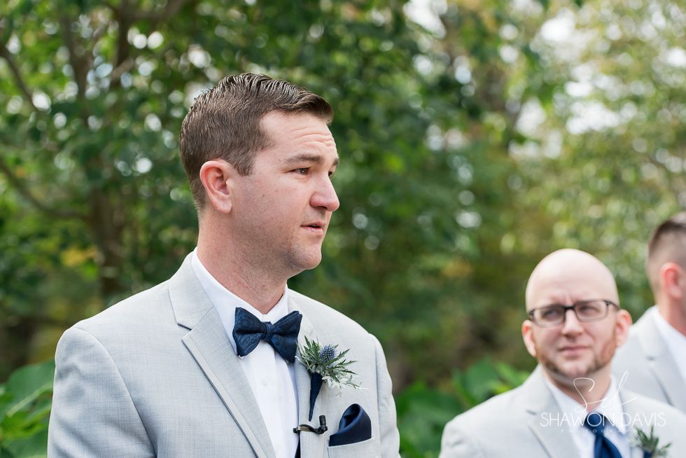 Man in light gray suit and navy bow tie outdoors at a wedding ceremony, with another man behind him.