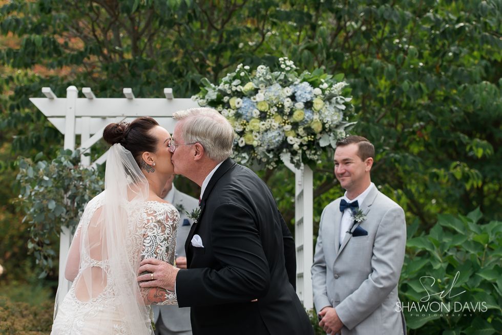 Bride and groom kiss at outdoor wedding ceremony, with officiant smiling nearby.