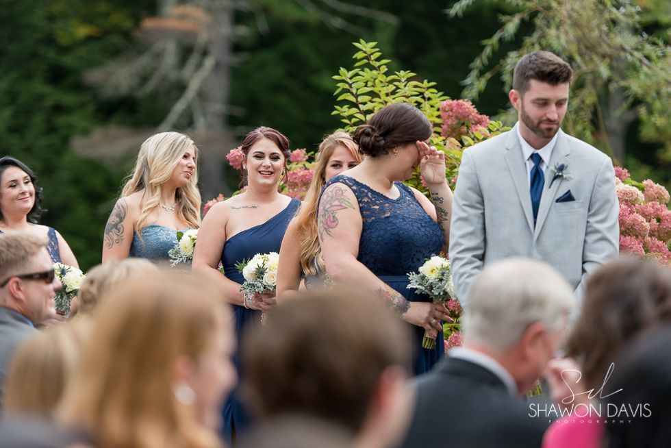 Guests in formal attire sit outdoors at a wedding, with a bride and groom standing among them.