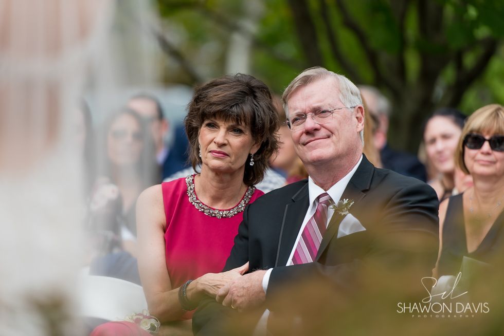 Two people seated outdoors at a formal event, one in a pink dress and one in a suit, smiling slightly.