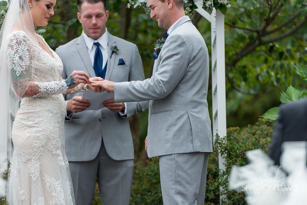 Wedding couple exchanging rings during an outdoor ceremony, officiant between them, in gray and white attire