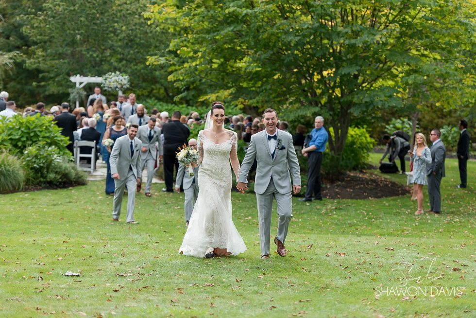 Newlyweds walking down a grassy aisle outdoors, surrounded by guests in a garden setting.