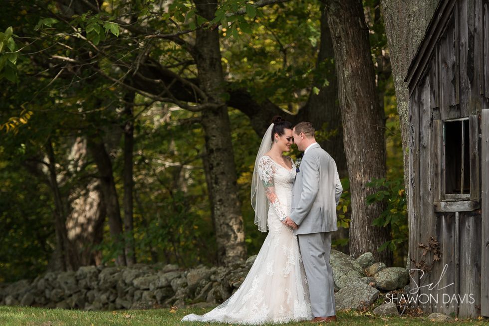 Bride and groom embracing in white attire beside a rustic stone wall in a wooded garden