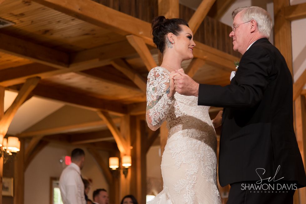Bride dances with an older man at a rustic indoor wedding reception, both smiling.