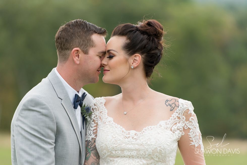 Bride and groom touching noses outdoors, wearing a light gray suit and white lace wedding dress.