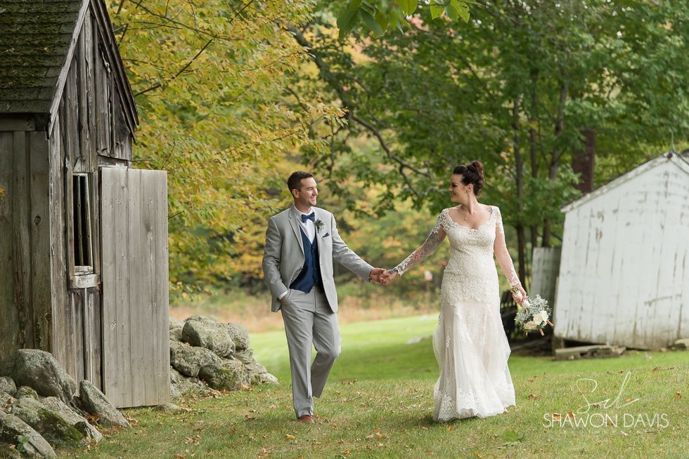 Couple holding hands outdoors by rustic barn and white shed in a green garden, smiling in wedding attire