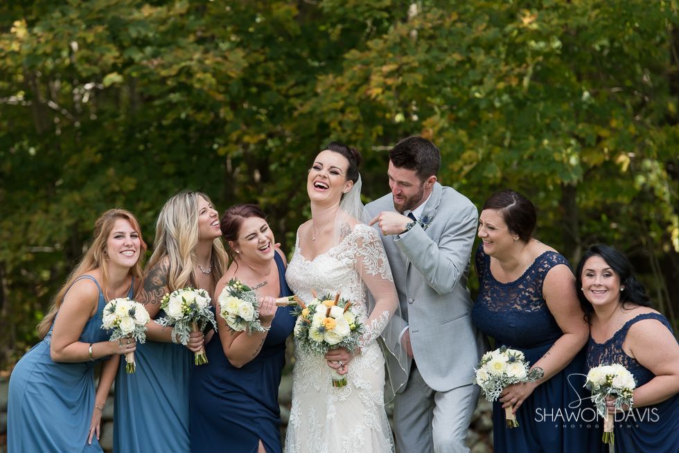 Bride and bridal party laughing outdoors in blue dresses and gray suit, holding bouquets