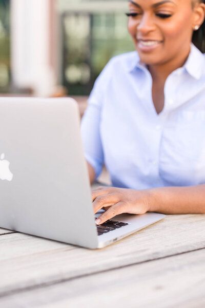 Smiling person using a silver laptop at an outdoor table, wearing a light blue shirt.