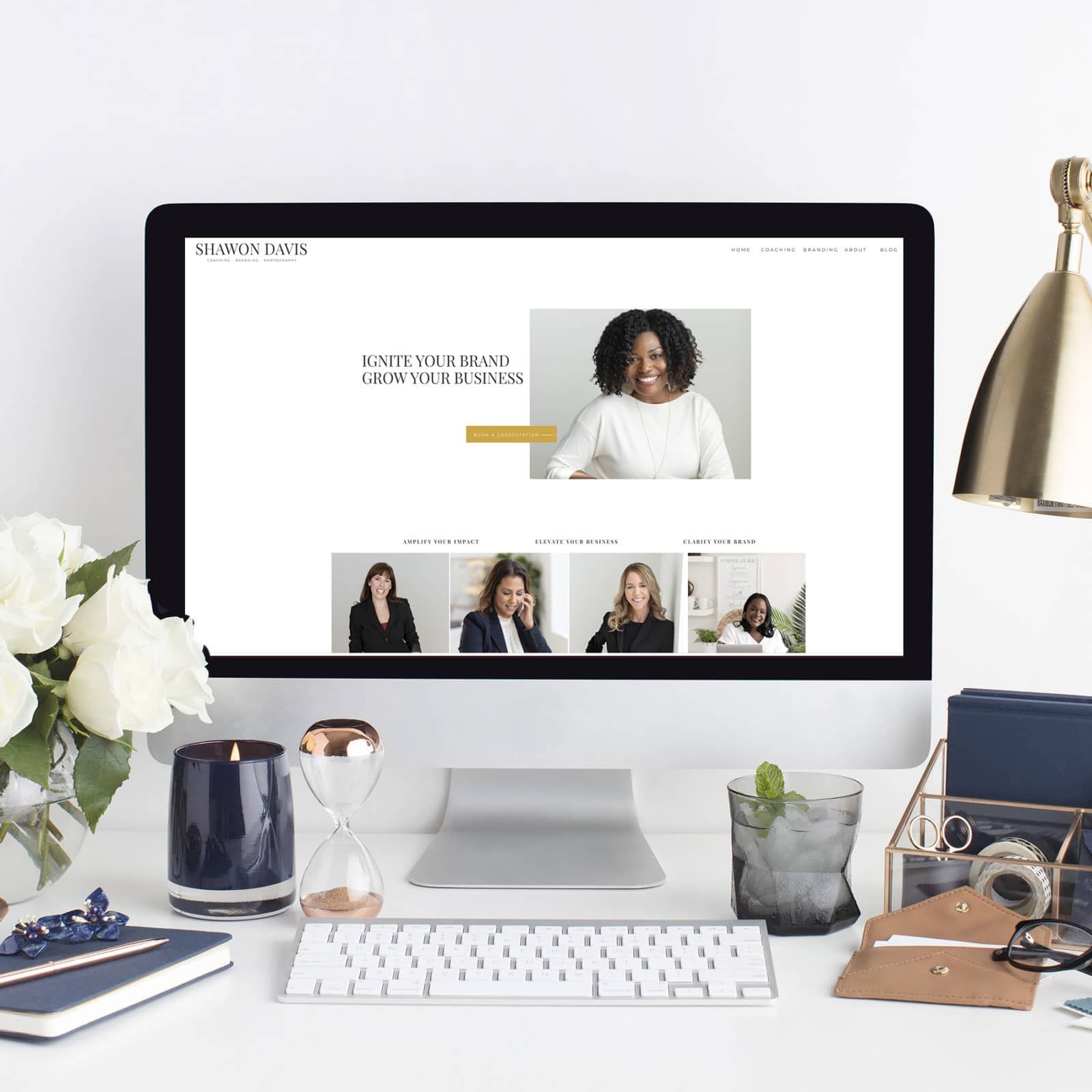 Desktop workspace with an iMac displaying a minimalist website on a white desk with flowers and decor