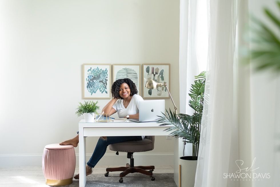 Woman smiling at a white desk in a bright home office with plants and framed wall art