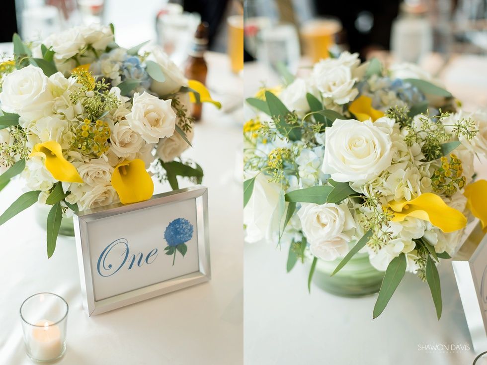 White and yellow floral centerpieces on a table with a “One” place card and teal accent