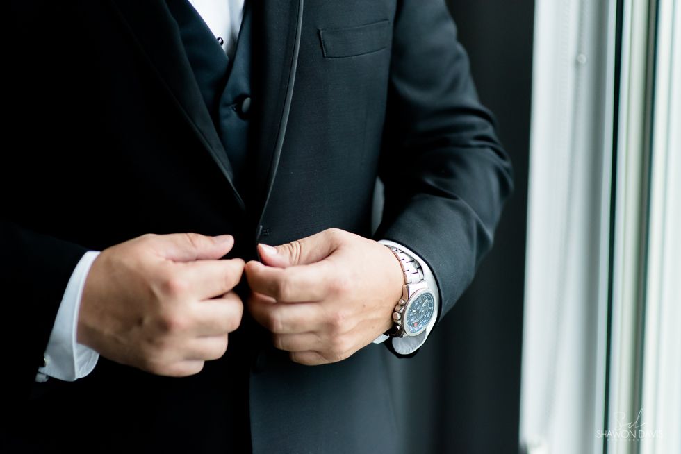 Person buttoning a black suit jacket near a window, wearing a silver watch.