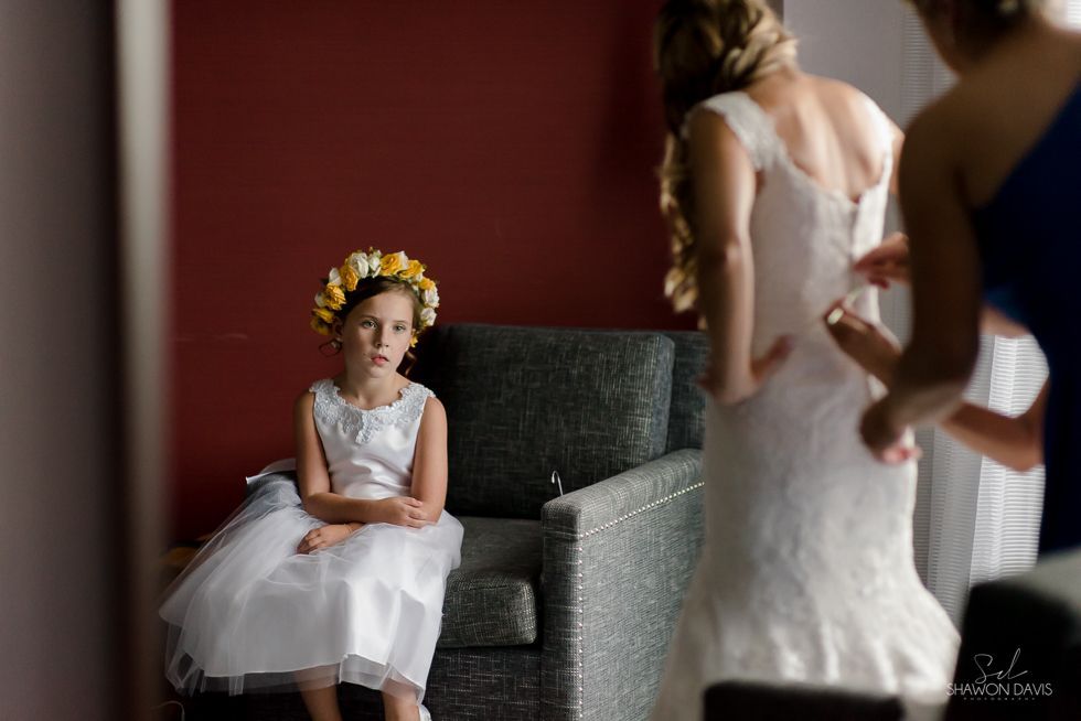 Flower-crowned child in a white dress seated on a chair, watching adults in wedding attire nearby.