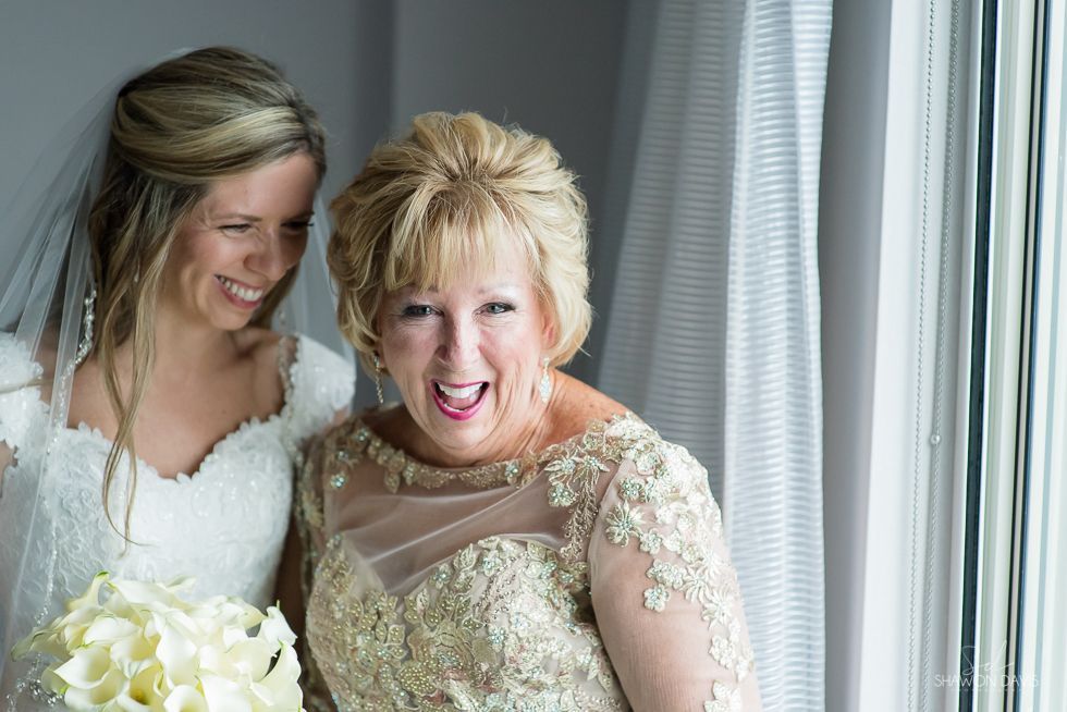 Bride and older woman smiling beside a window, both in white dresses and holding a bouquet