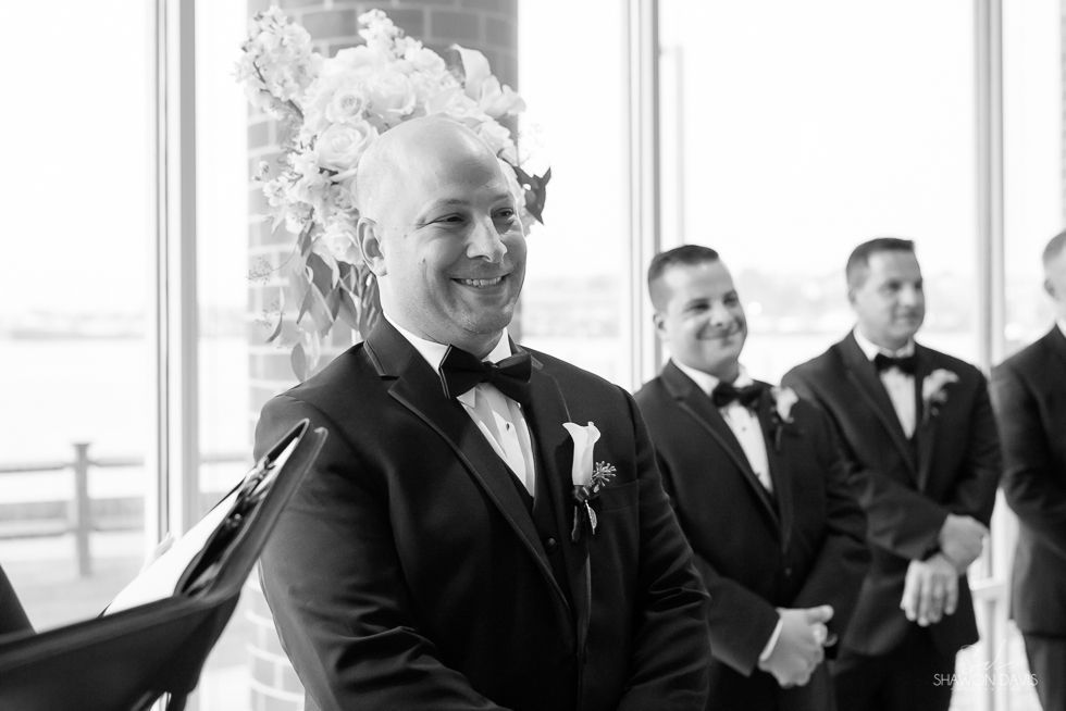 Smiling groom in tuxedo with boutonniere, standing indoors near two groomsmen in suits