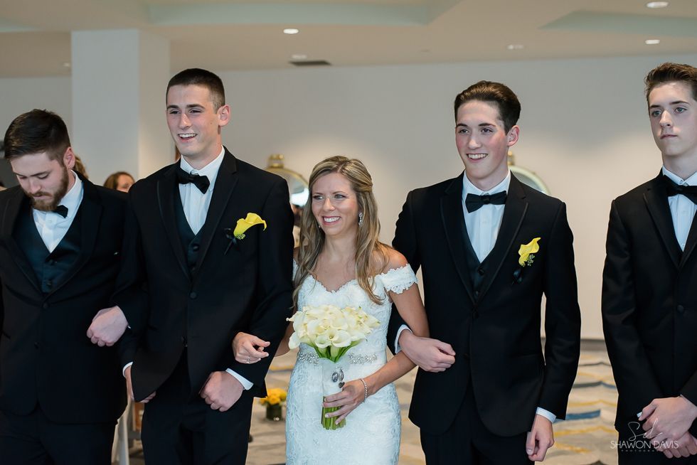 Wedding party walking indoors, with the bride in a white dress holding yellow flowers and groomsmen in black tuxedos.